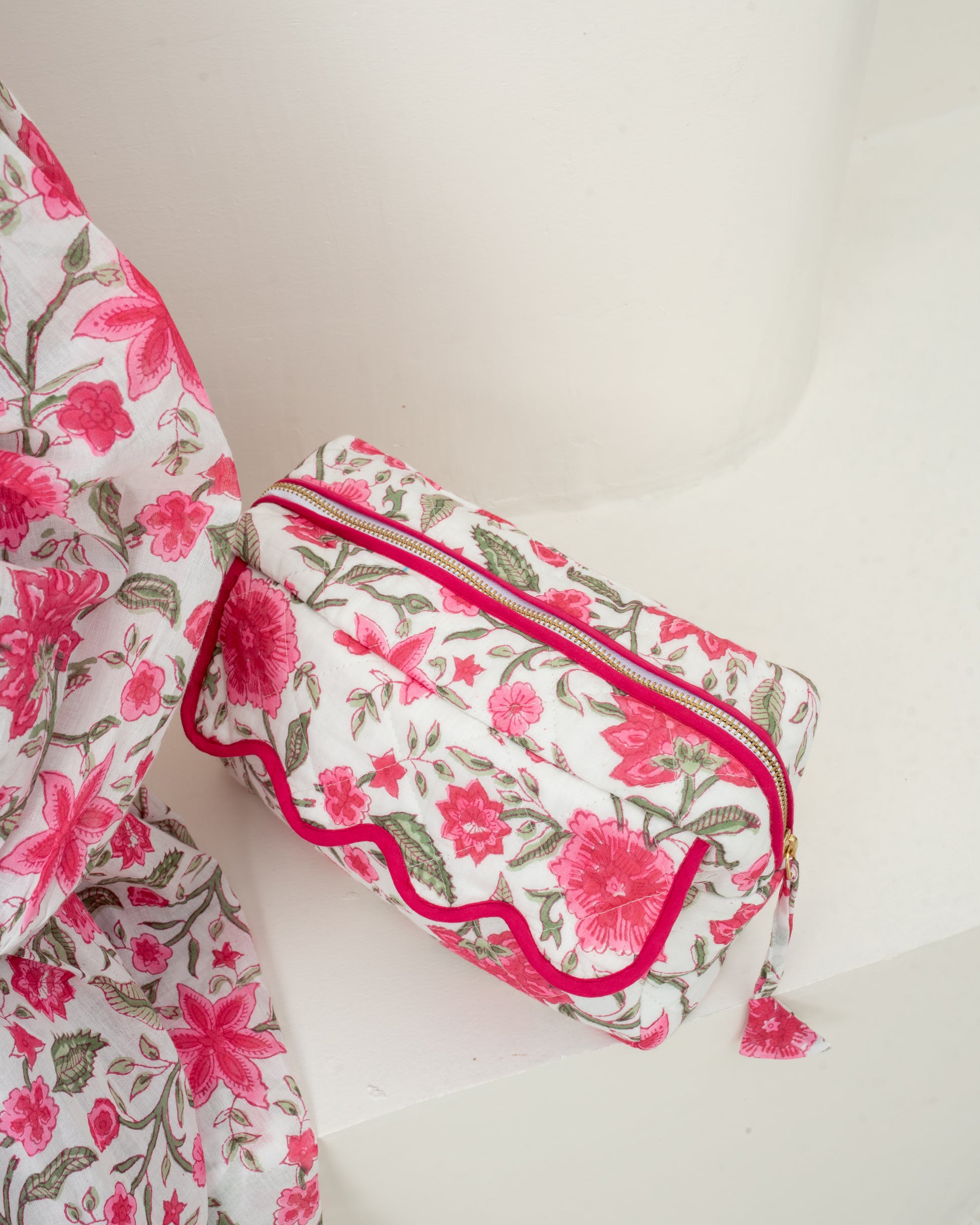 A small rectangular zip pouch with a pink and green floral print on a white background, accented with bright pink trim and a matching tassel zipper pull, shown next to a matching floral fabric piece on a white surface.