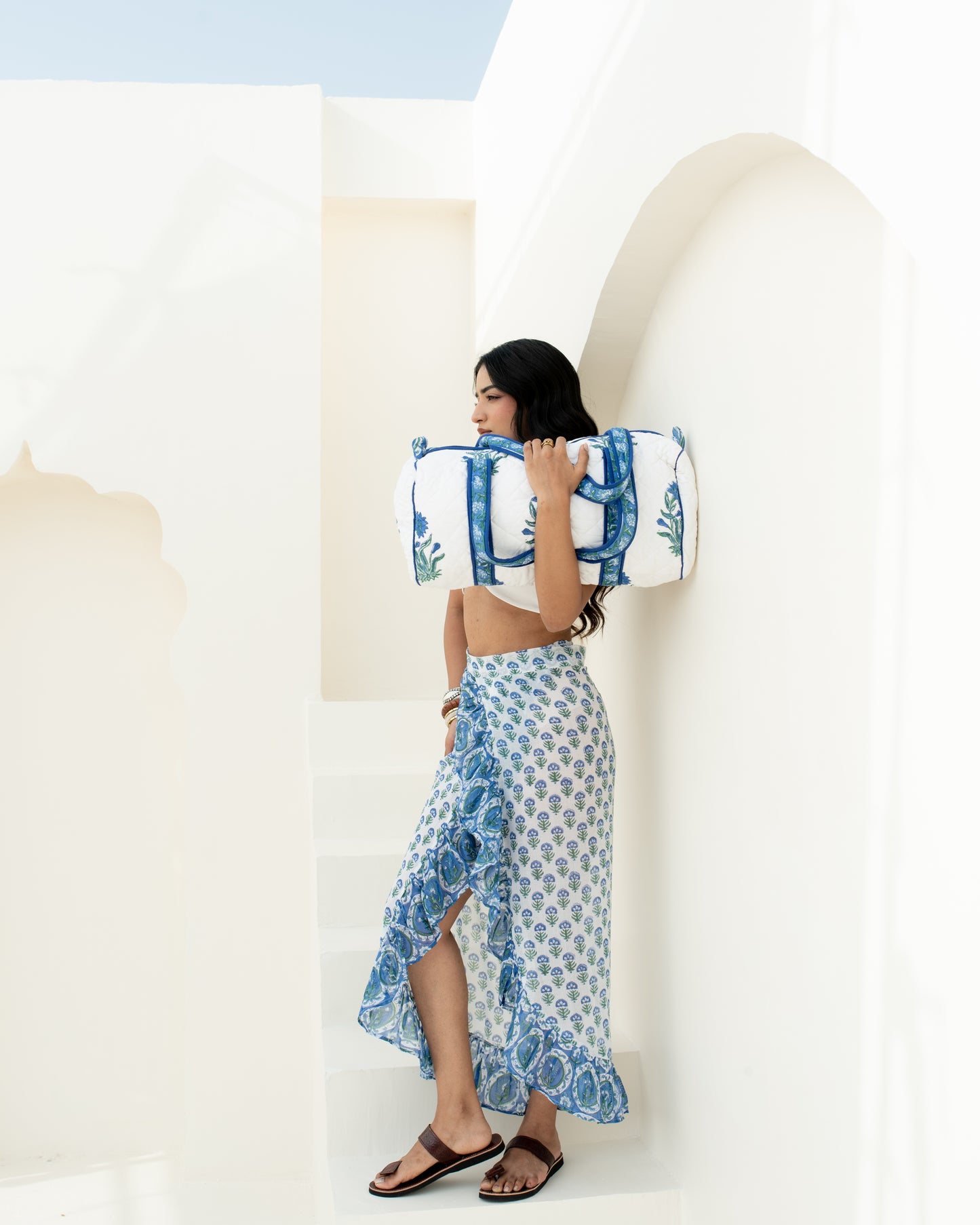 A woman stands on white architectural steps carrying a large white and blue patterned duffel bag over her shoulder, wearing a matching blue and white patterned wrap skirt and white crop top.