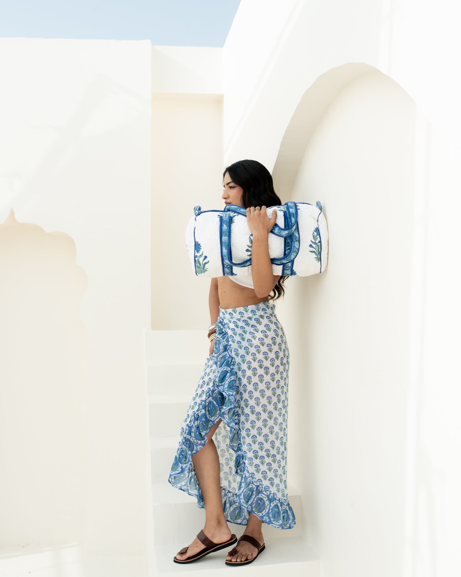 A woman stands on white architectural steps carrying a large white and blue patterned duffel bag over her shoulder, wearing a matching blue and white patterned wrap skirt and white crop top.