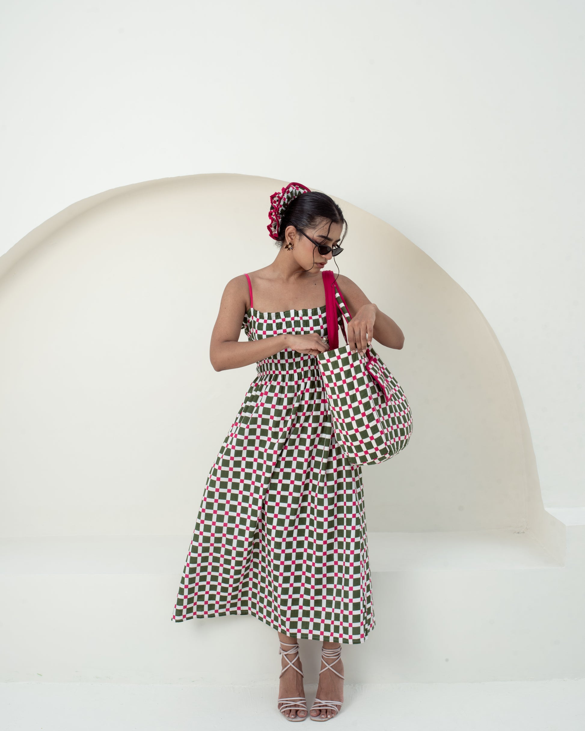 A woman modeling a sleeveless midi dress with a red, green, and black geometric pattern, holding a matching patterned handbag against a minimalist white background with a curved architectural element.