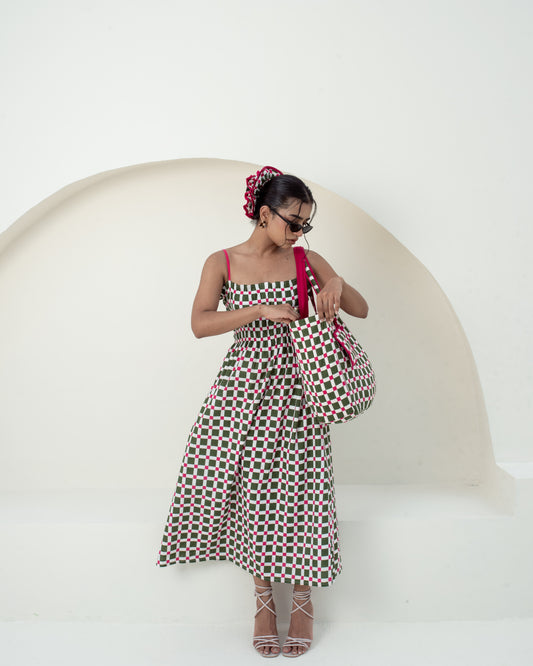 A woman modeling a sleeveless midi dress with a red, green, and black geometric pattern, holding a matching patterned handbag against a minimalist white background with a curved architectural element.