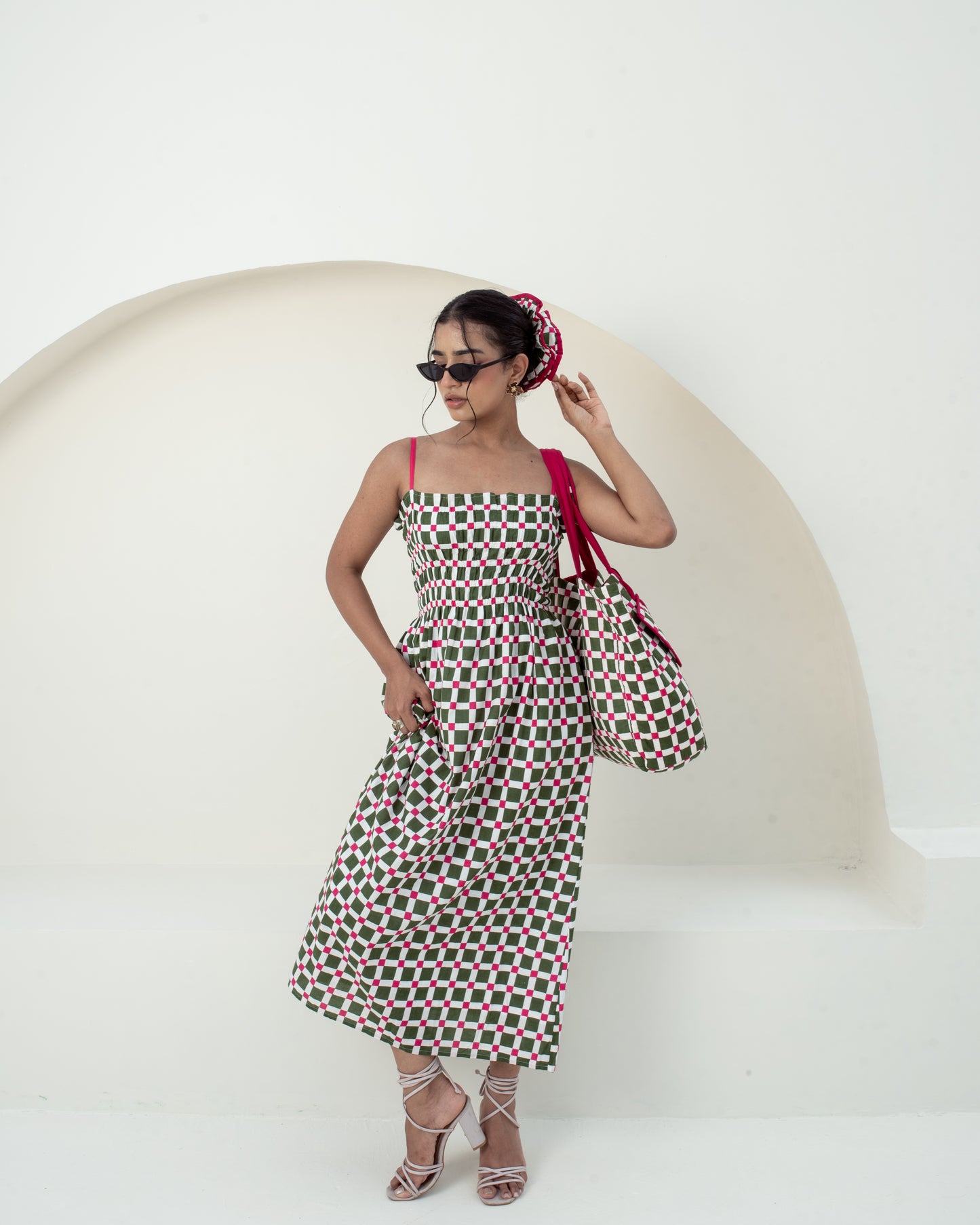 A woman models a sleeveless midi dress with a green, pink, and white geometric pattern, styled with a matching pink headscarf, sunglasses, and a large pink shoulder bag against a minimalist white background with a curved architectural detail.