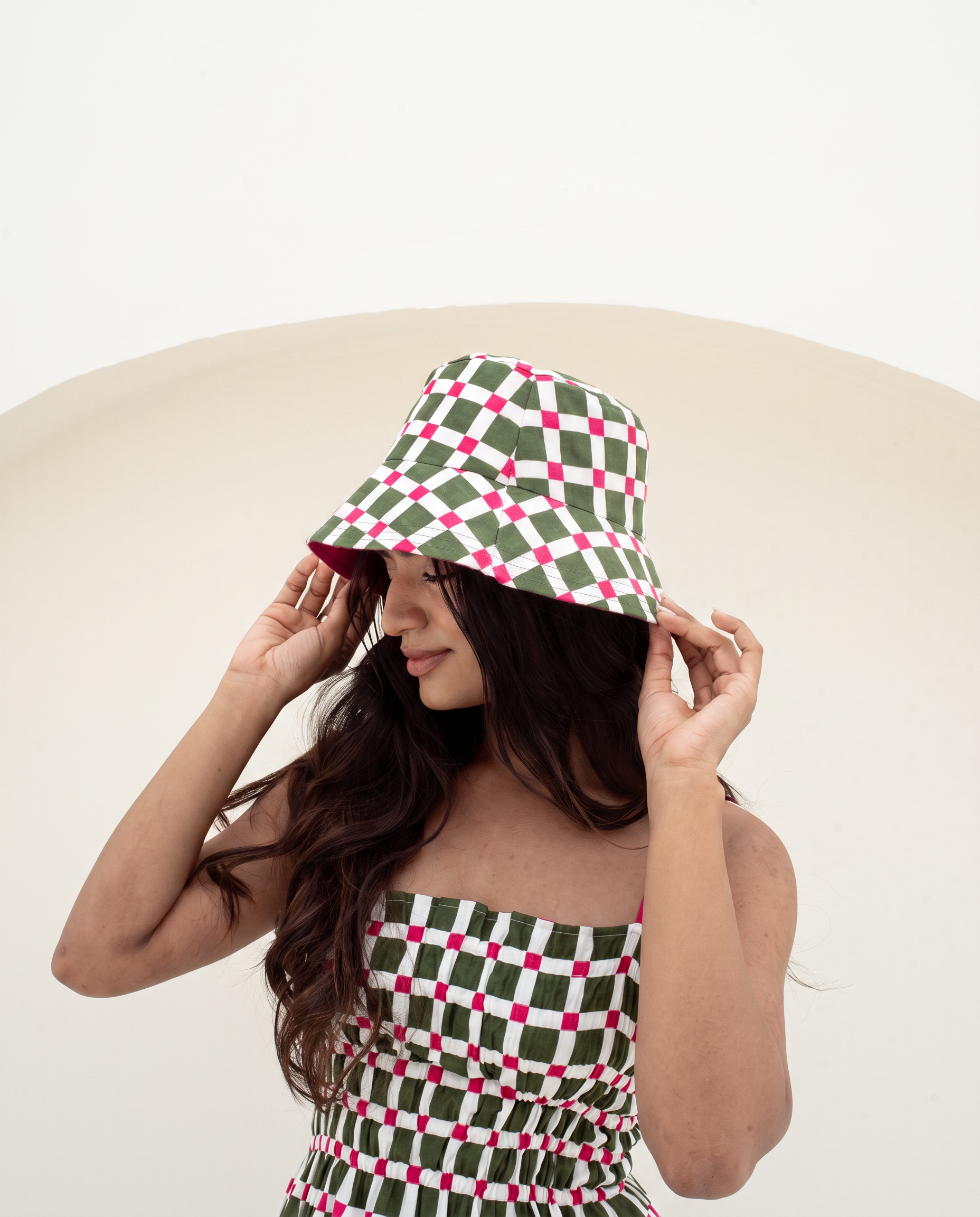 A woman wearing a green, white, and pink checkered bucket hat and a matching checkered strapless top, posing against a minimalist beige and white background.