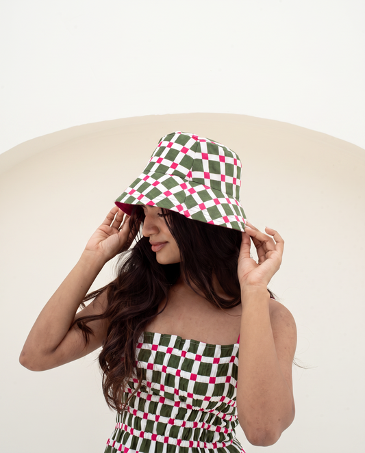 A woman wearing a green, white, and pink checkered bucket hat and a matching checkered strapless top, posing against a minimalist beige and white background.